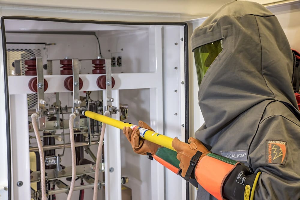 A substation operator in full arc-flash PPE using a hot stick and high-voltage detector to test a busbar